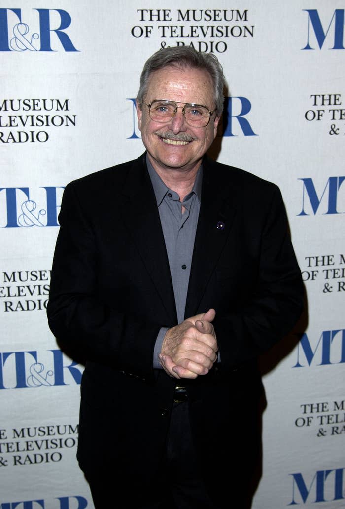 Bill Daniels smiles at a red carpet event. Bill's wearing a simple suit and eyeglasses