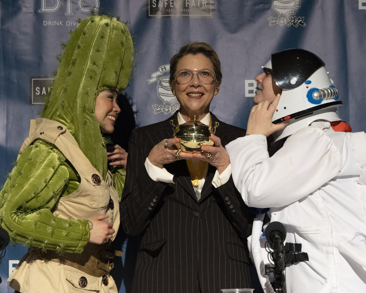 Annette Bening onstage holding the pudding pot award, flanked by two members of Harvard University’s Hasty Pudding Theatricals who were dressed as a cactus and an astronaut.