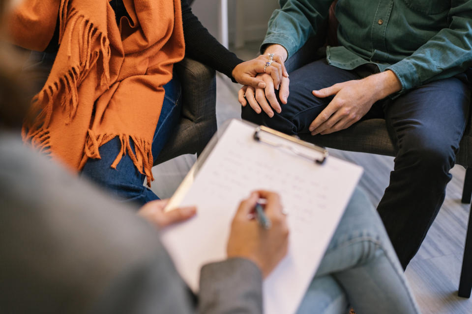 Three people in a meeting, one taking notes on a clipboard, another gesturing with hands