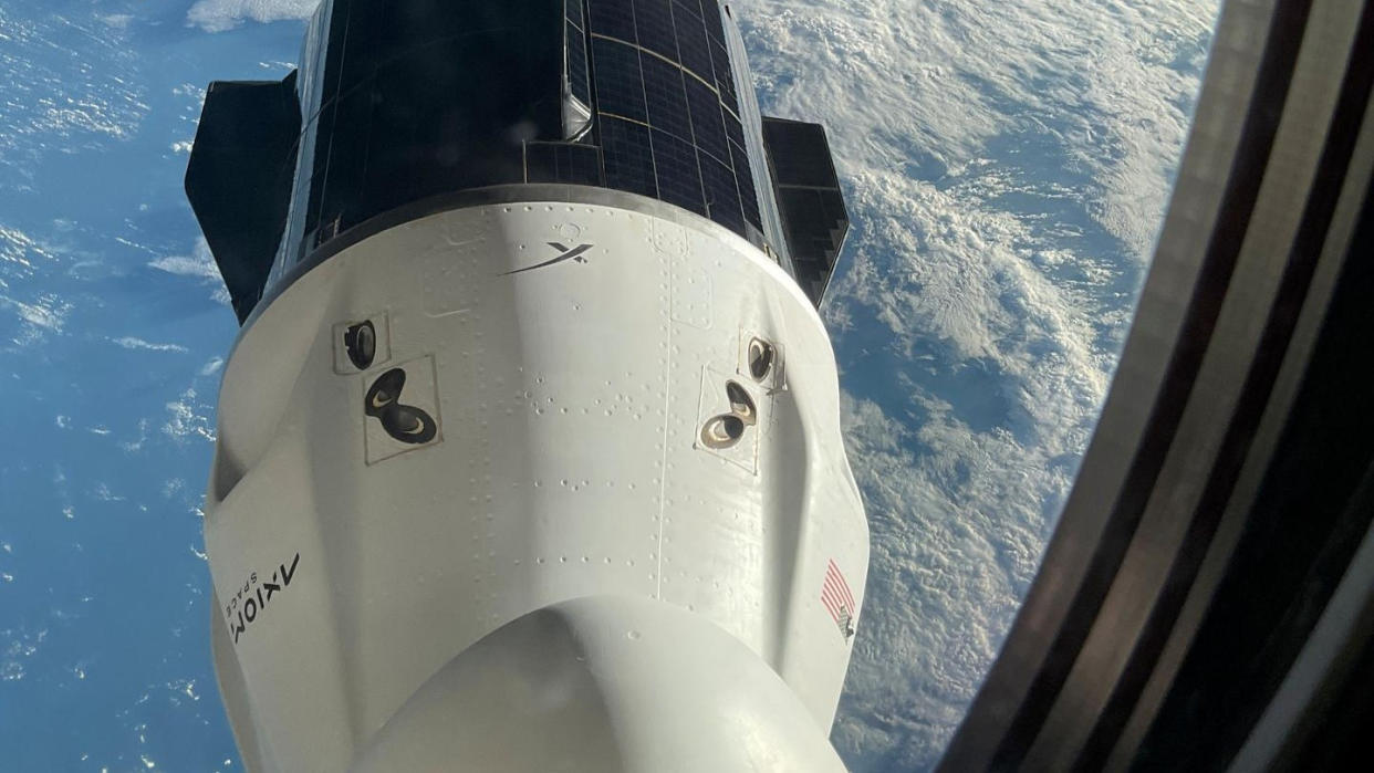  Through a rounded window looking downward, a white space capsule is seen with dark solar panels lining its aft trunk. Below, the blue of Earth, with a wash of white clouds. 