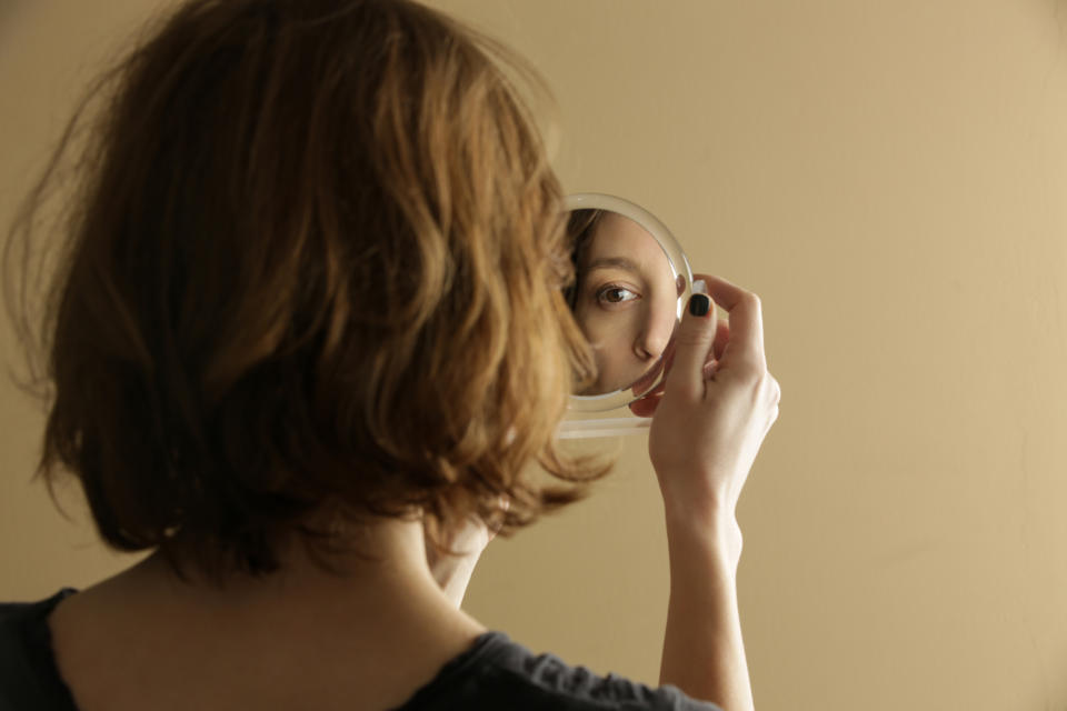 A person with shoulder-length hair is looking at their reflection in a handheld mirror, revealing only one eye