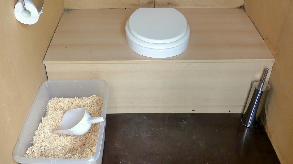 toilet seat on wooden bench with bucket of saw dust, toilet brush, and toilet paper
