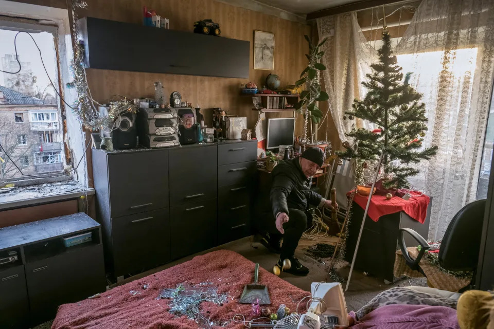 A local resident, kneeling by his Christmas tree in his living room, removes shards of glass from broken windows in his flat in a residential building.
