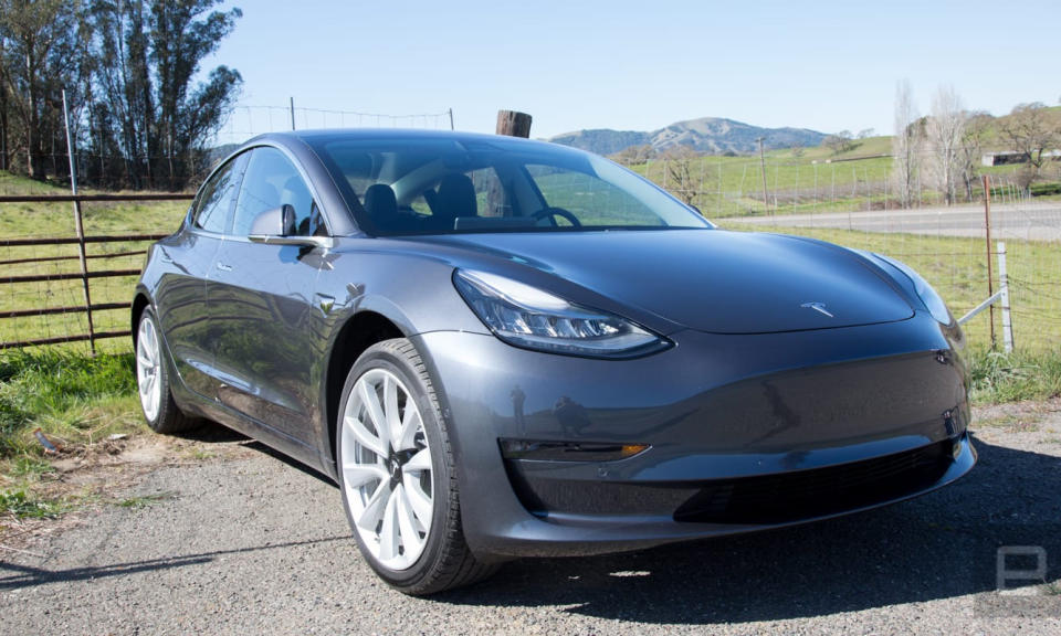 Photo of the Tesla Model 3 sitting outdoors next to a field. Green grass, trees and hills are visible in the distance.