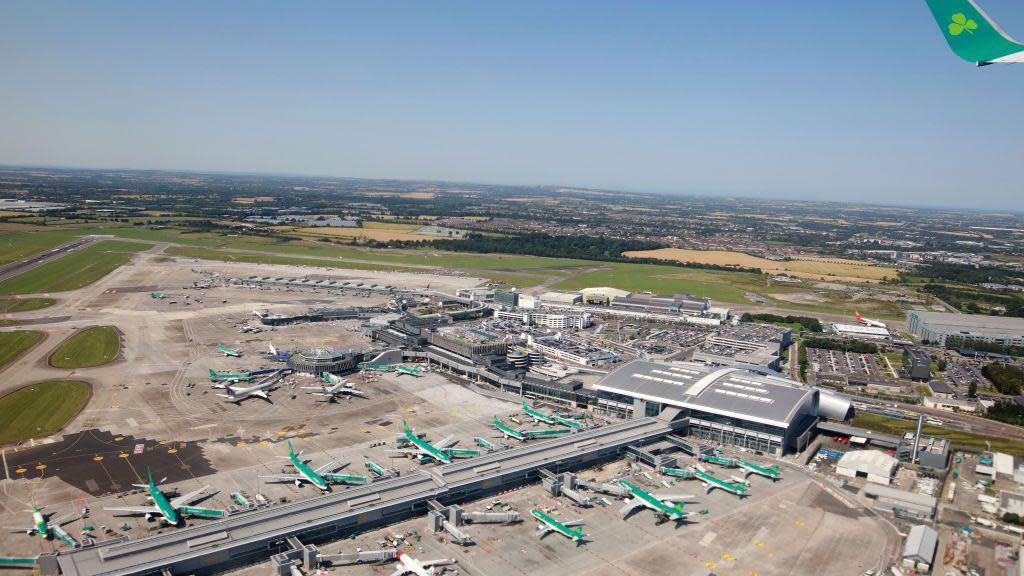 An aerial view of Dublin Airport on a bright day. Several Aer Lingus aircraft can be seen parked at stands outside the airport building.