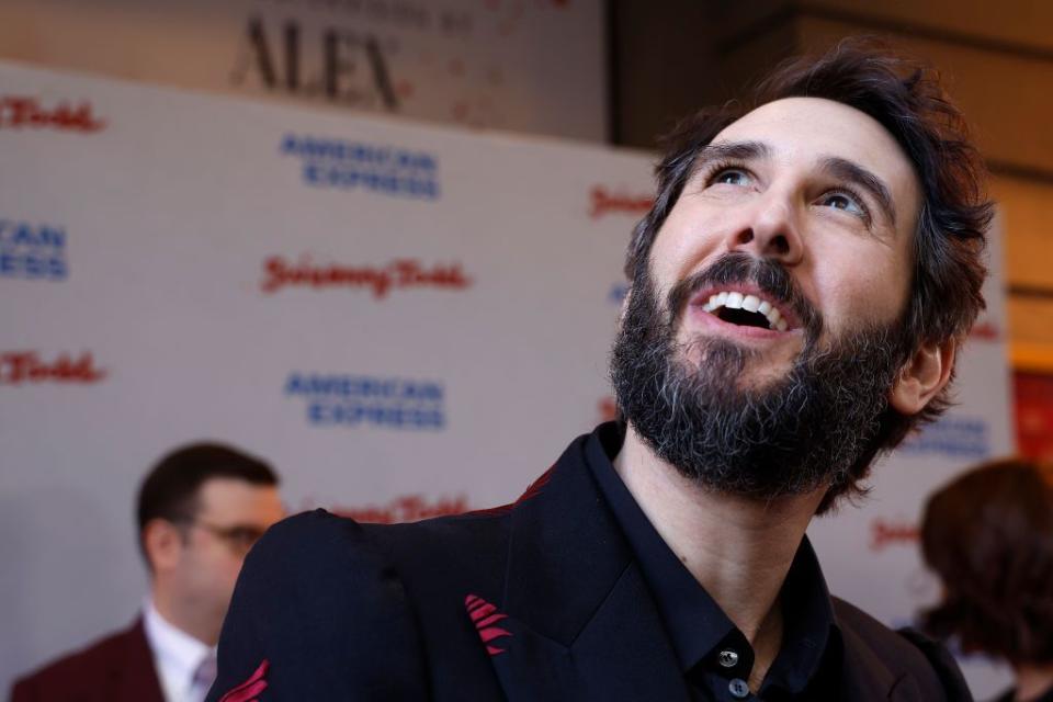 josh groban smiles and looks up while wearing a black and red suit, with a sweeney todd advertisement behind him