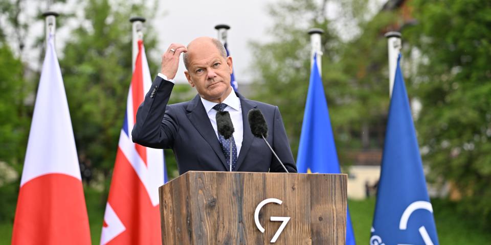 Germany's Chancellor Olaf Scholz scratching his head at a podium in front of G7 flags.
