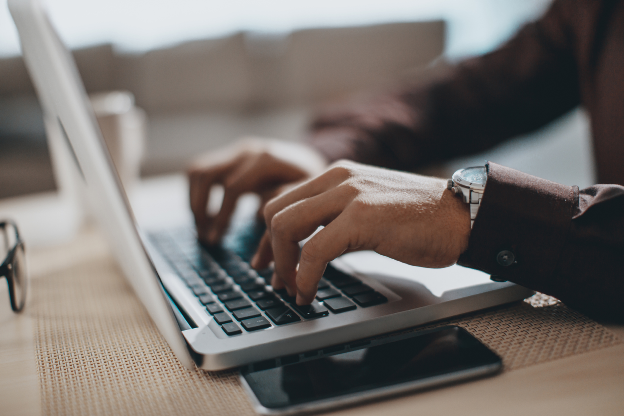 Closeup of two man's hands typing on a MacBook with smartphone next to it on a desk at home, with glasses and a paper coffee cup, with a blurred background of a couch and a wall