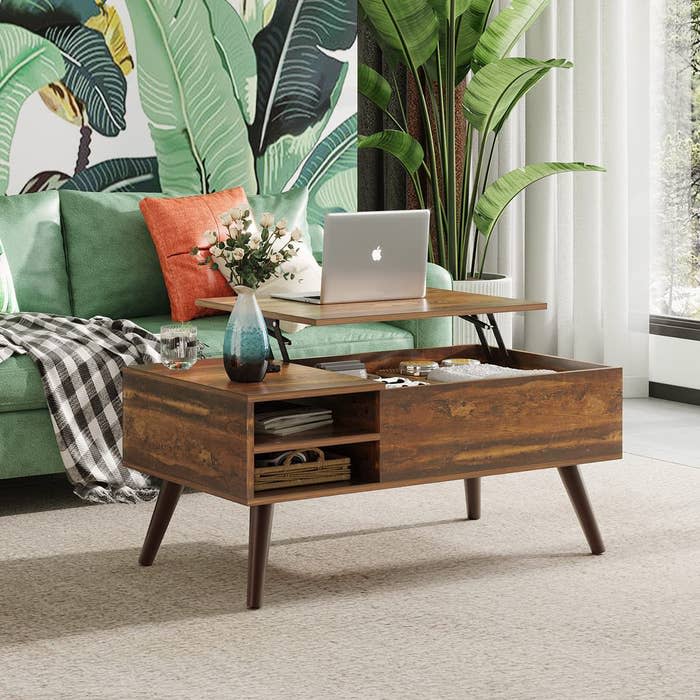 Lift-top wooden coffee table with hidden storage and decorative items, in a modern living room with a green sofa, plant wallpaper, and a laptop on the table