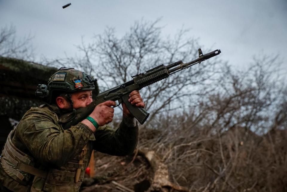 A Ukrainian serviceman in camouflage gear fires an AK-74 with bare trees and a grey sky behind him