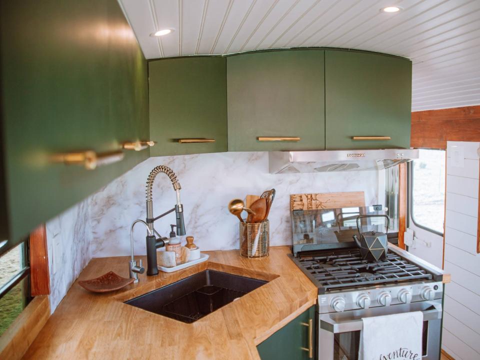 The kitchen inside the school bus, with green cabinets, a white blacksplash, and wood counters