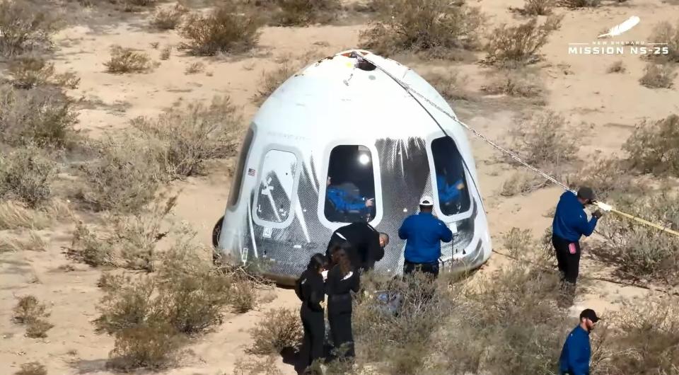 Técnicos vestidos con monos negros caminan alrededor de una cápsula blanca en forma de cono en el suelo del desierto.