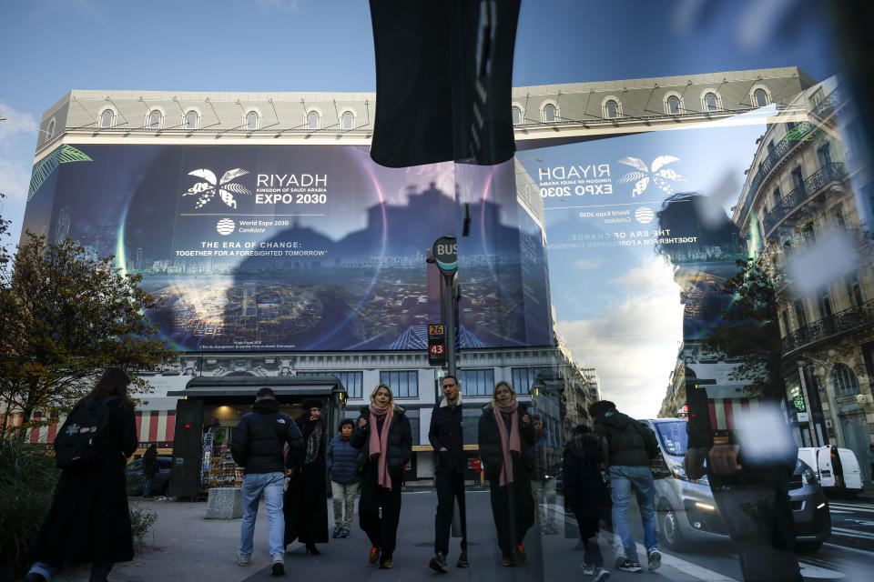 People walk by a building under renovation protected with a canvas decorated with a commercial promoting the Riyadh Expo 2030, Tuesday, Nov. 28, 2023 in Paris. In a high-profile showdown, Rome, Busan and Riyadh are the top contenders to become the host city of the 2030 World Expo, with the organizing body choosing the winner on Tuesday. (AP Photo/Thomas Padilla)