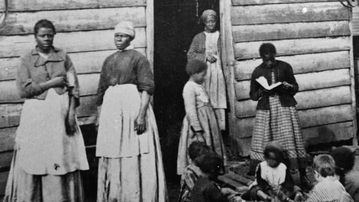 A group of women and children, presumably enslaved, sit and stand around the doorway of a rough wooden cabin, Southern United States, mid 19th Century. One girl reads a book to the group of sitting children. Black people were enslaved by slavemasters for profit.