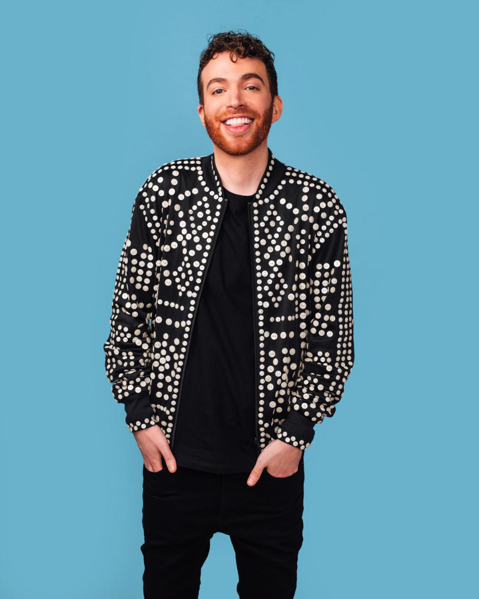 Josh smiles widely as they poses with both hands their pocket and rocks a polka dot jacket