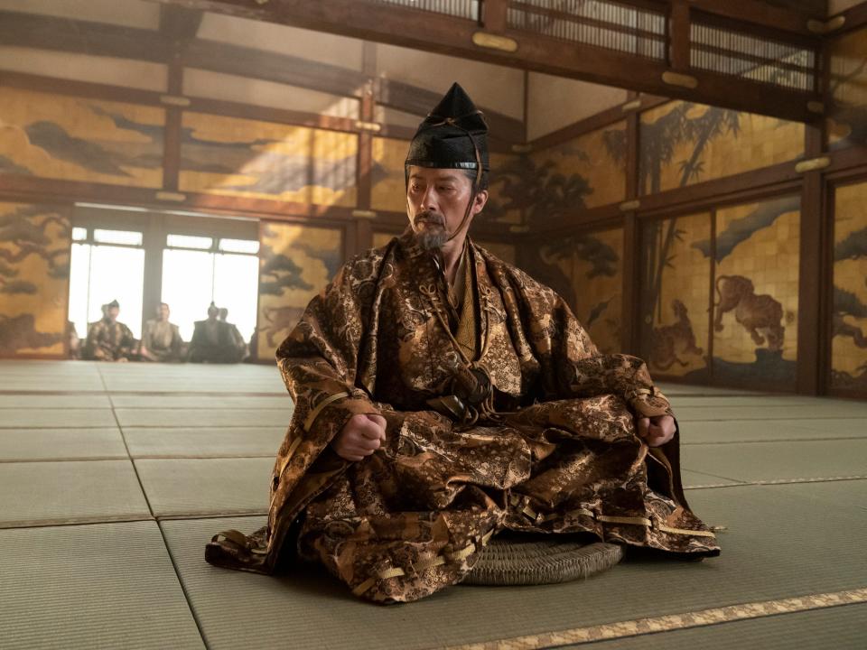hiroyuki sanada as yoshii toranaga, a man wearing ornate golden clothing and an eboshi headpiece, sitting alone in a room crosslegged