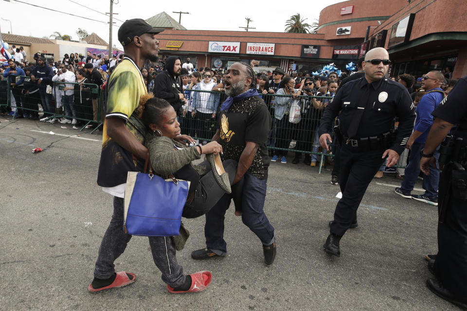 Two men carry a woman injured in a stampede as people gather to watch a hearse carrying the casket of slain rapper Nipsey Hussle Thursday, April 11, 2019, in Los Angeles. The 25-mile procession traveled through the streets of South Los Angeles after a memorial service, including a trip past Hussle's clothing store, The Marathon, rear, where he was gunned down March 31. (AP Photo/Jae C. Hong)