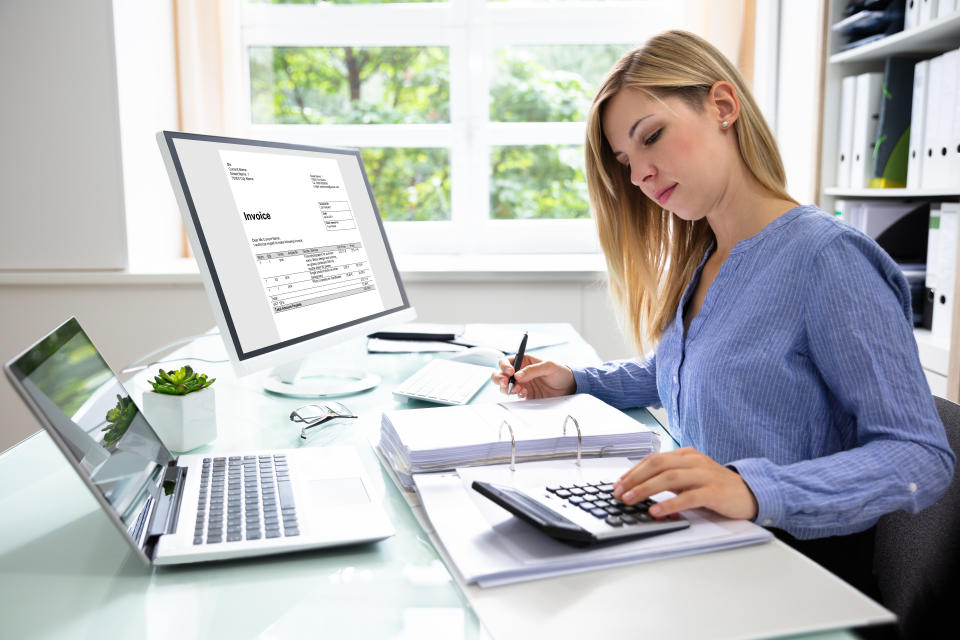 Young businesswoman using a calculator to work out tax relief while sitting at a desk with a computer and laptop.