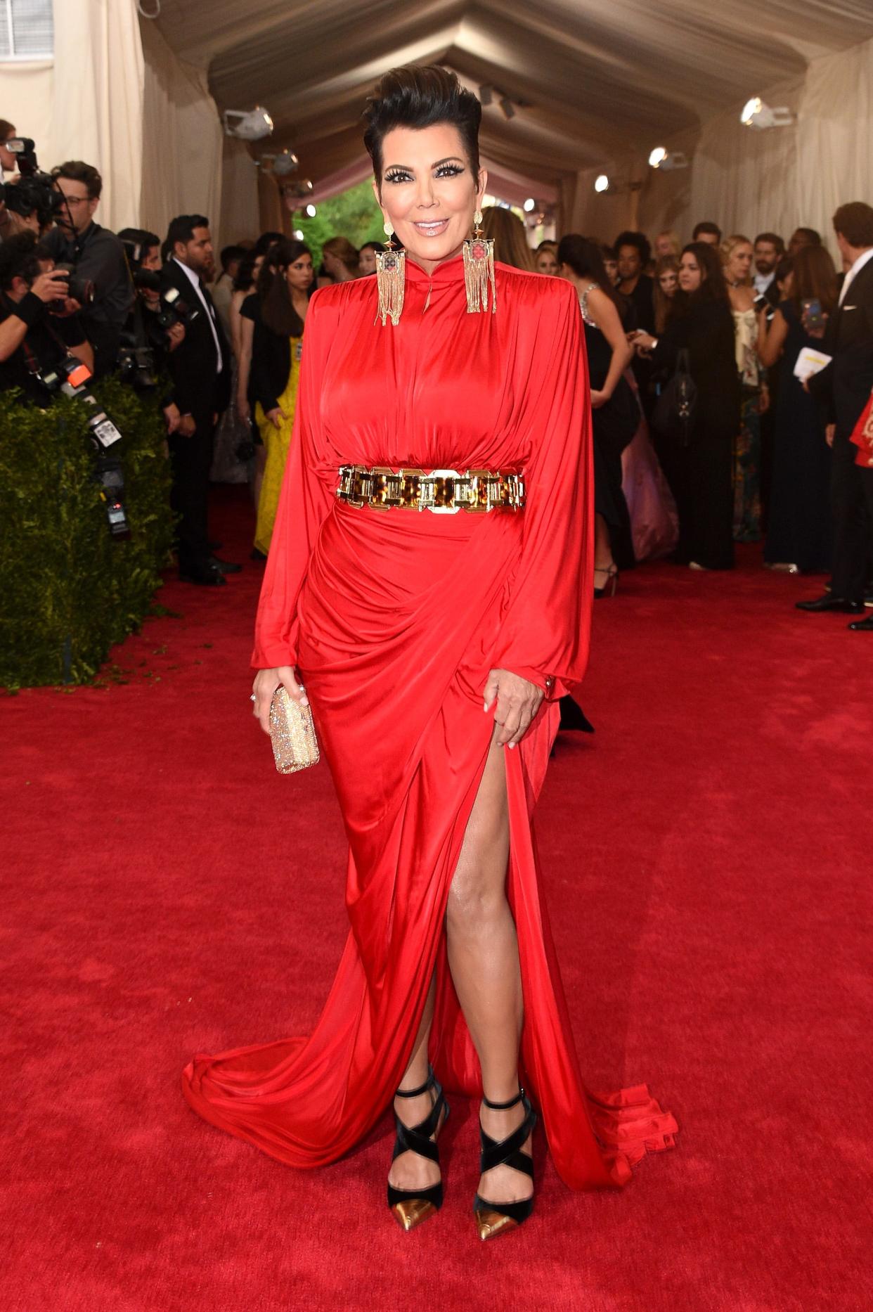 Kris Jenner attends the 2015 Met Gala in a red gown with a thigh-high slit that she accessorized with a gold belt, dangling gold earrings, and black strappy heels with gold pointed toes.