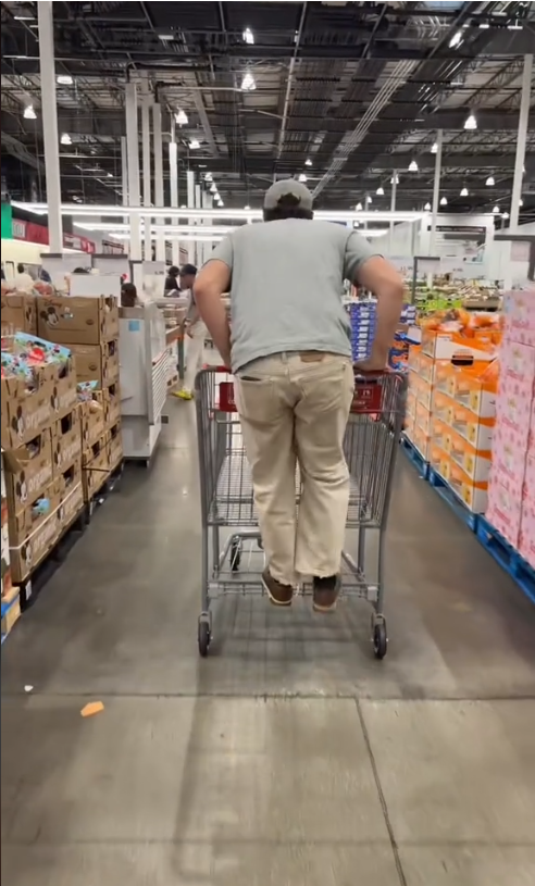 Person riding a shopping cart through a large store aisle, surrounded by boxes and produce displays