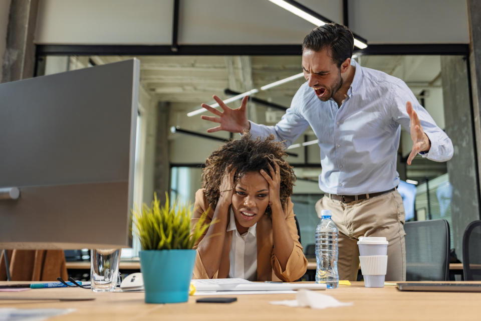 Man angrily yells at a frustrated woman sitting at an office desk with paperwork and a computer monitor. The scene appears tense and stressful