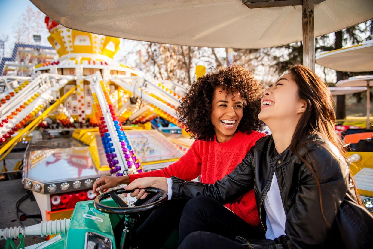 Two young woman laughing while riding a rotating circular amusement park ride, one woman's hand is on a wheel, with colorful center of ride and trees without leaves in the background