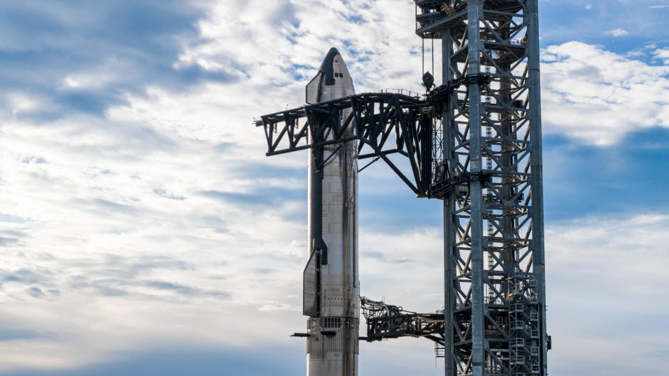 closeup photo of a shiny silver conical spacecraft sitting atop a first-stage booster by the metallic arms of a launch tower.