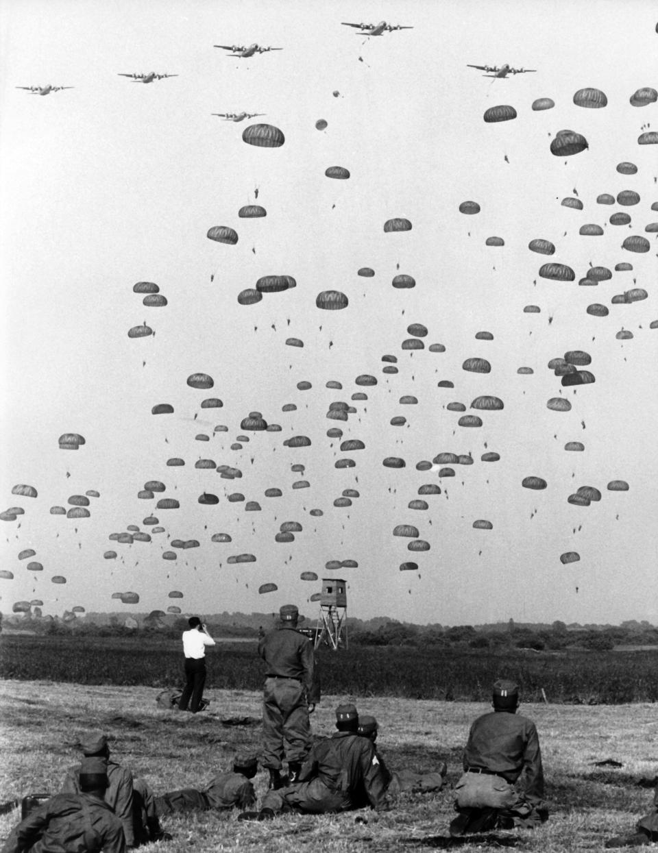 A black-and-white photo of soldiers staring out at a horizon filled with dozens of parachuting soldiers floating down into a field.