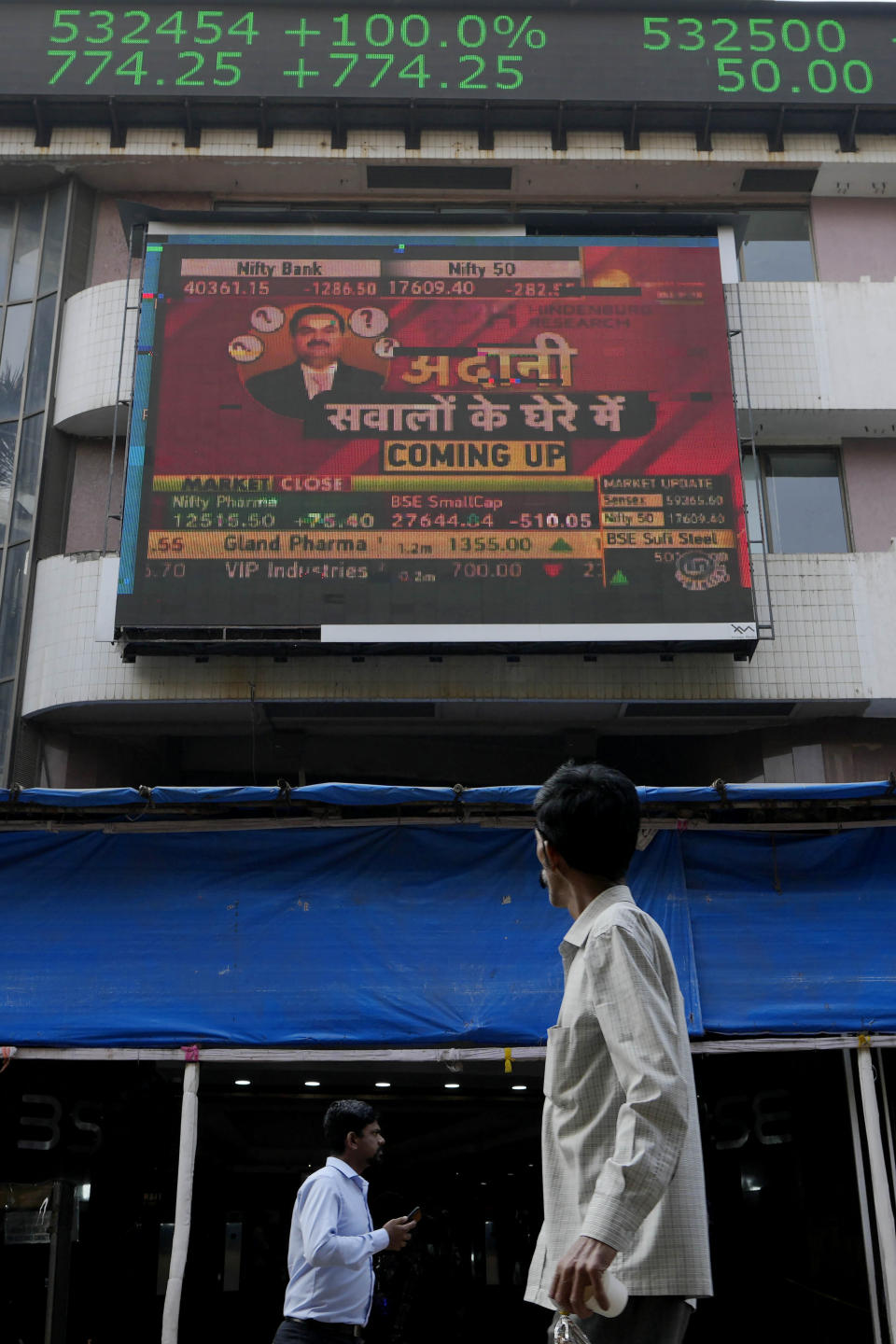 People walk past an electronic display featuring news about Adani Group outside the Bombay Stock Exchange building in Mumbai, India, Friday, Jan. 27, 2023. India’s Adani Group launched a share offering for retail investors Friday as it mulled taking legal action against U.S.-based short-selling firm Hindenburg Research over a report that led investors to dump its shares, with some stocks in the group falling up to 20% on Friday. (AP Photo/Rajanish Kakade)