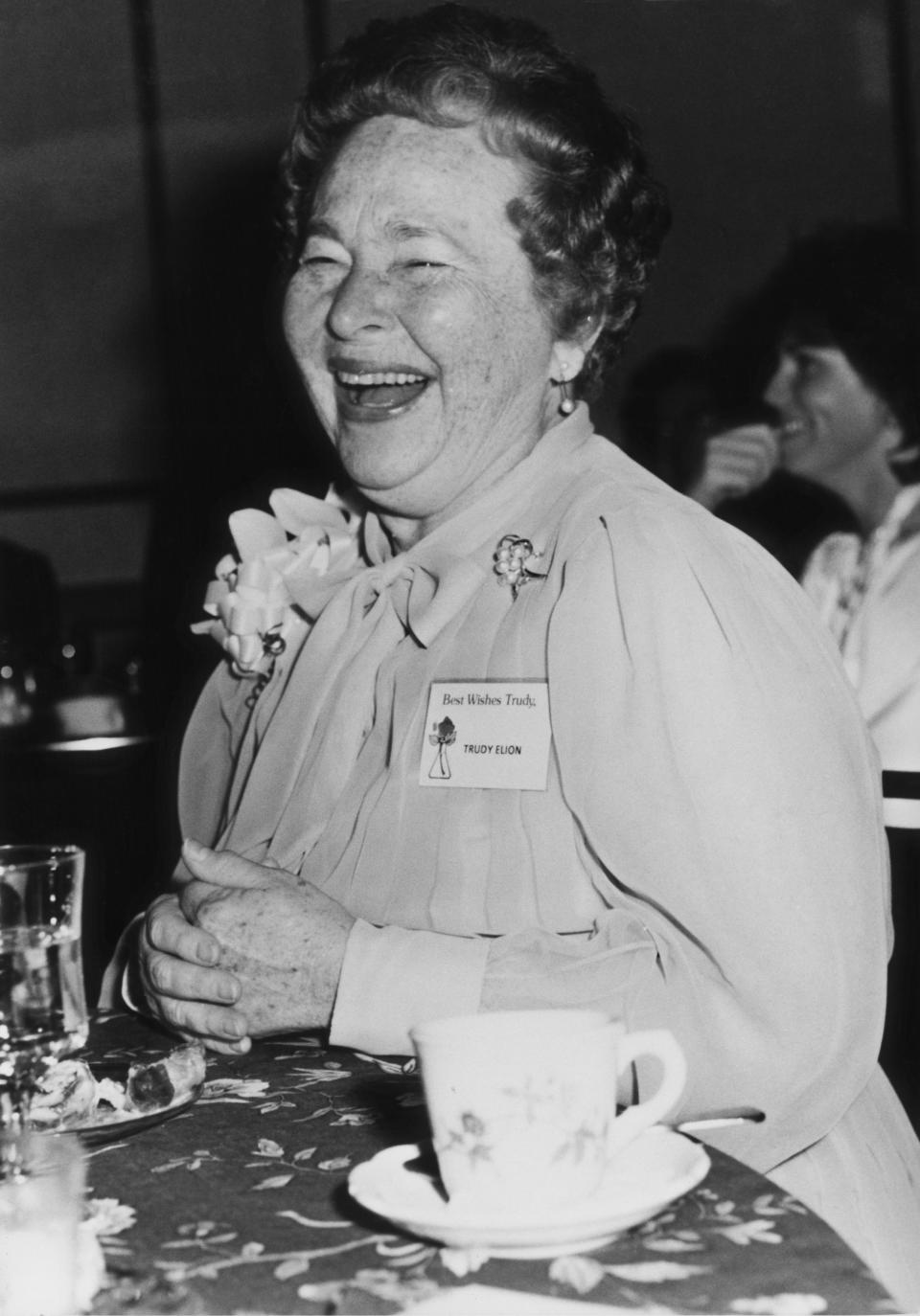 Gertrude Elion laughing, sitting at a table with a floral tablecloth and a teacup and saucer in front of her