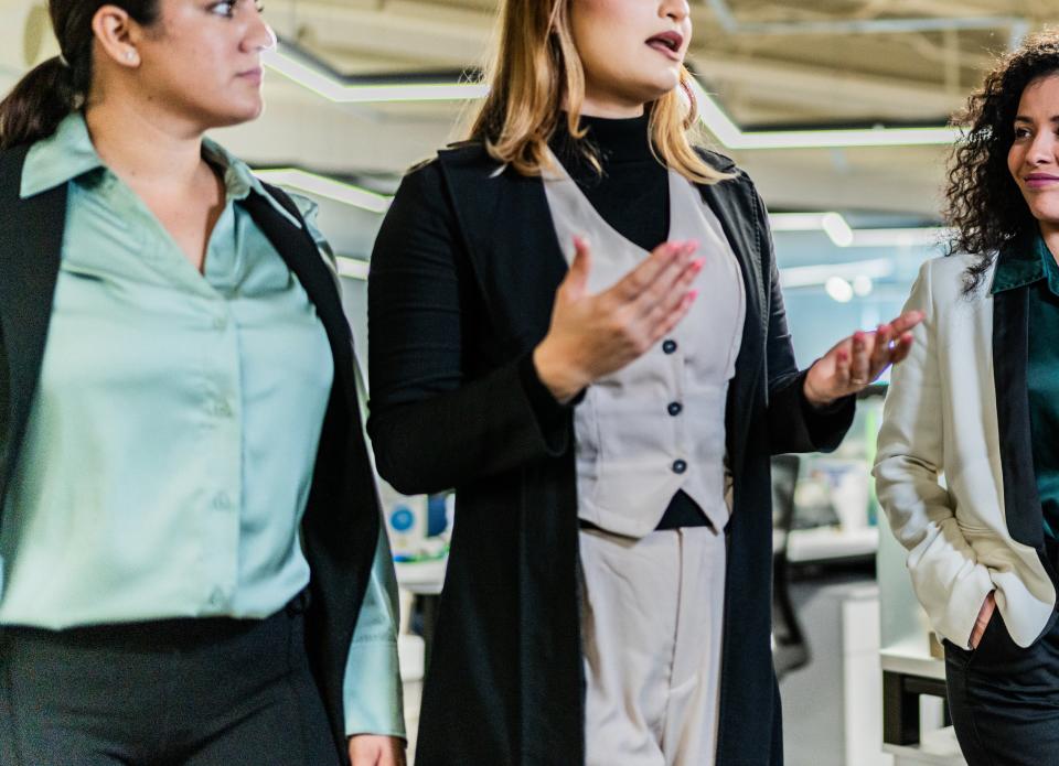 Three women in professional attire discuss while walking through a modern office space