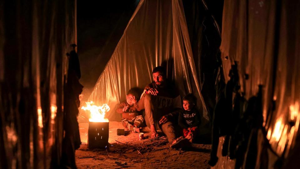 PHOTO: A man sits with children by a fire outside one of the tents housing Palestinians displaced by the conflict in Gaza between Israel and the Palestinian Hamas movement, in Rafah in the southern Gaza Strip, Dec. 18, 2023. (Mahmud Hams/AFP via Getty Images)