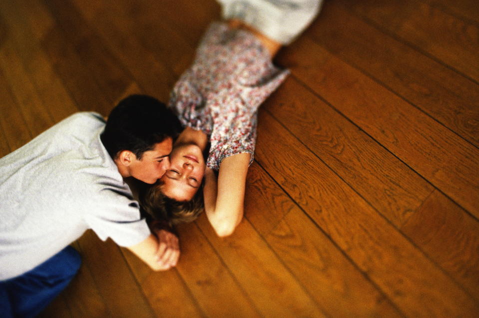 Two people lying on a wooden floor, embracing and smiling at each other