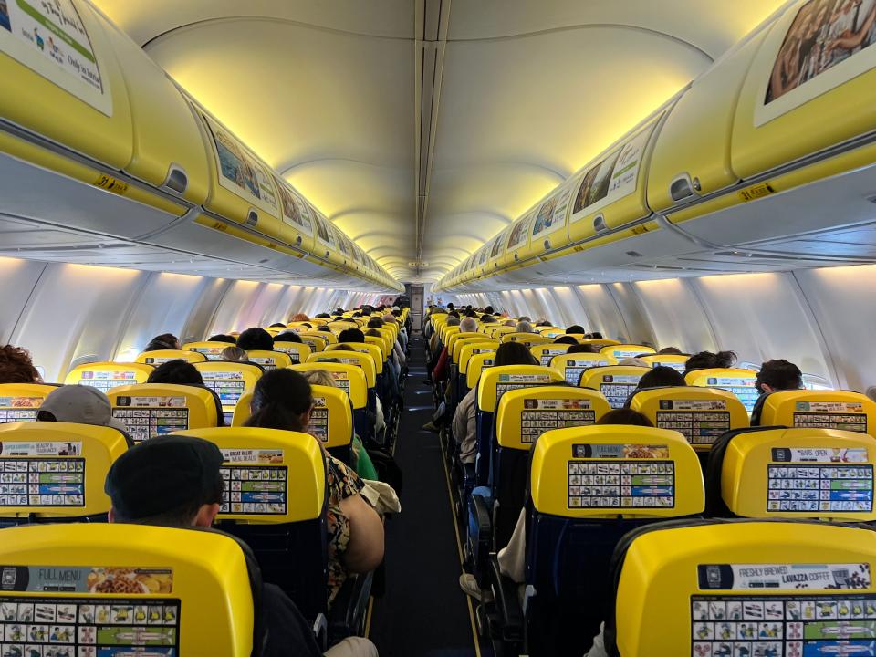 The cabin of a Ryanair Boeing 737-800 in its yellow and blue color scheme with commercials on the overhead bins, as viewed from the middle of the aisle.
