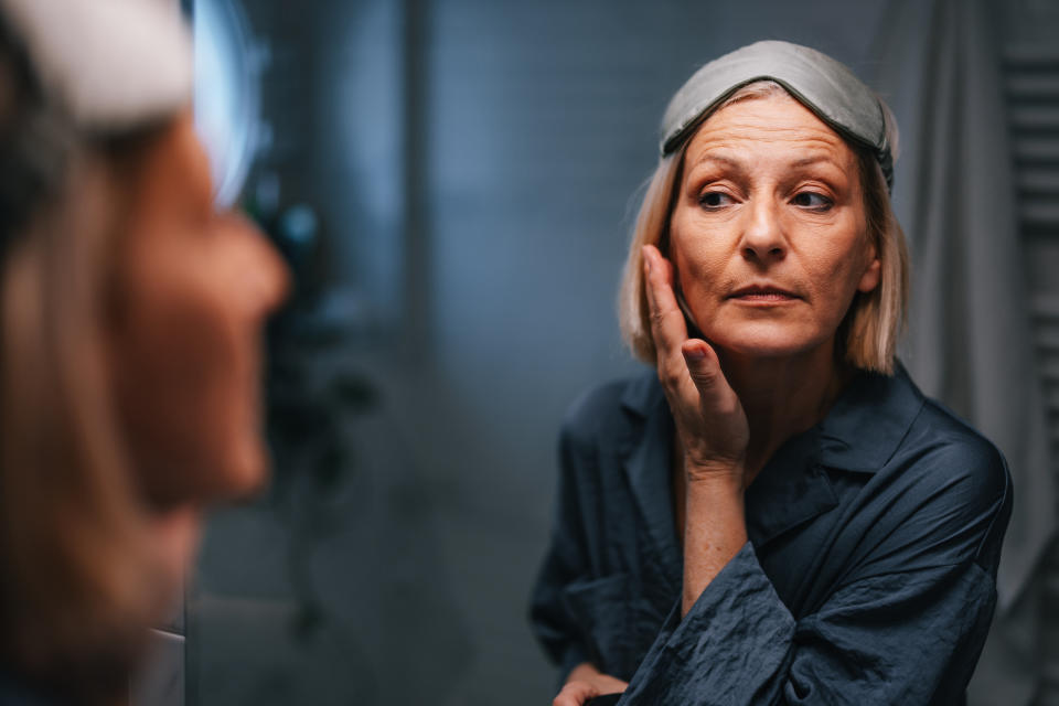 A woman in a sleep mask and robe examines her face in the mirror with a thoughtful expression