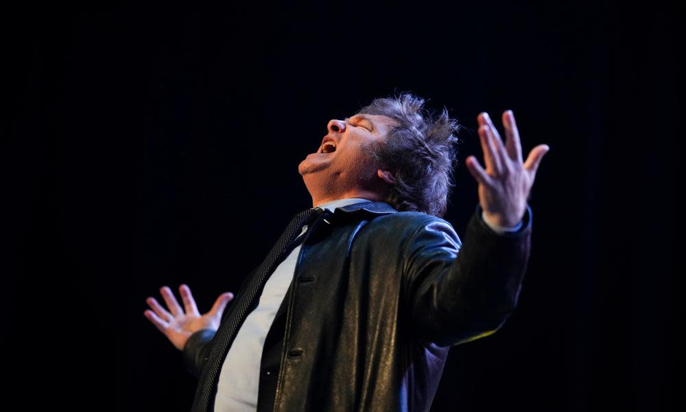 A white man, head thrown back, eyes closed, mouth open, arms and fingers outstretched, bathed in light in an otherwise dark frame. Wears a T-shirt and black jacket.