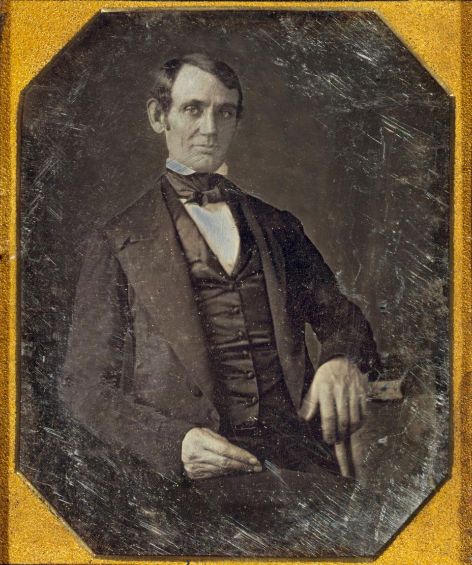 Abraham Lincoln, in a formal portrait, sits while wearing a dark suit and a bow tie, with his hand resting on a chair