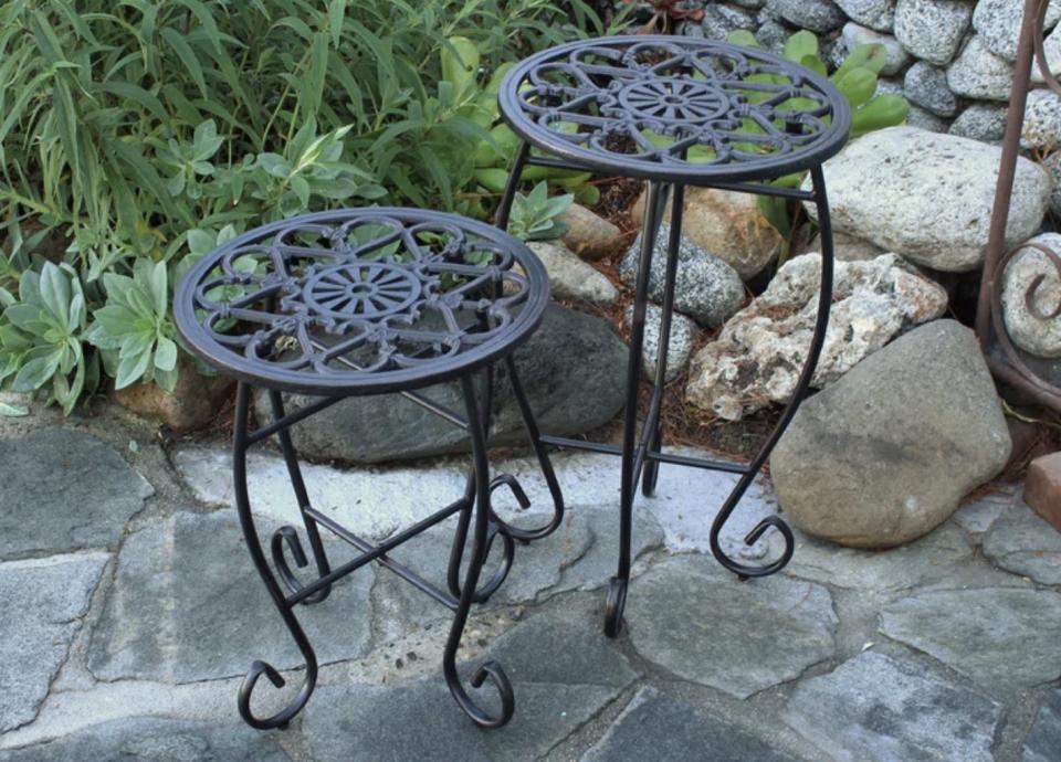 Two round metal garden stools with ornate patterns, displayed on a stone patio with plants in the background