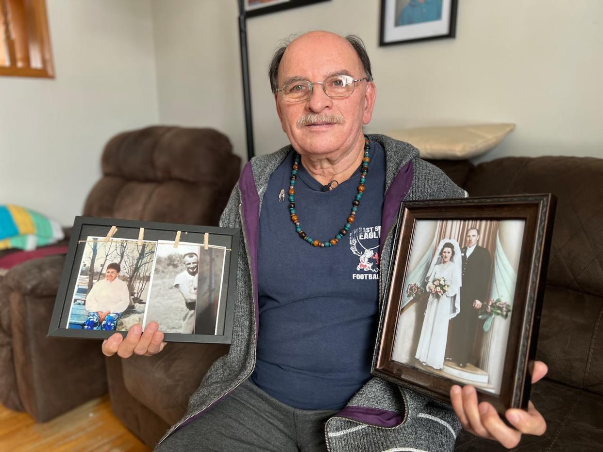 Edward Ambrose holds up two picture frames in this file photo from 2023. The picture in his left hand is a wedding photo of his 'bonded parents,' he calls them. The pictures in his right hand are of his biological parents, whom he never got to meet. (Gary Solilak/CBC - image credit)