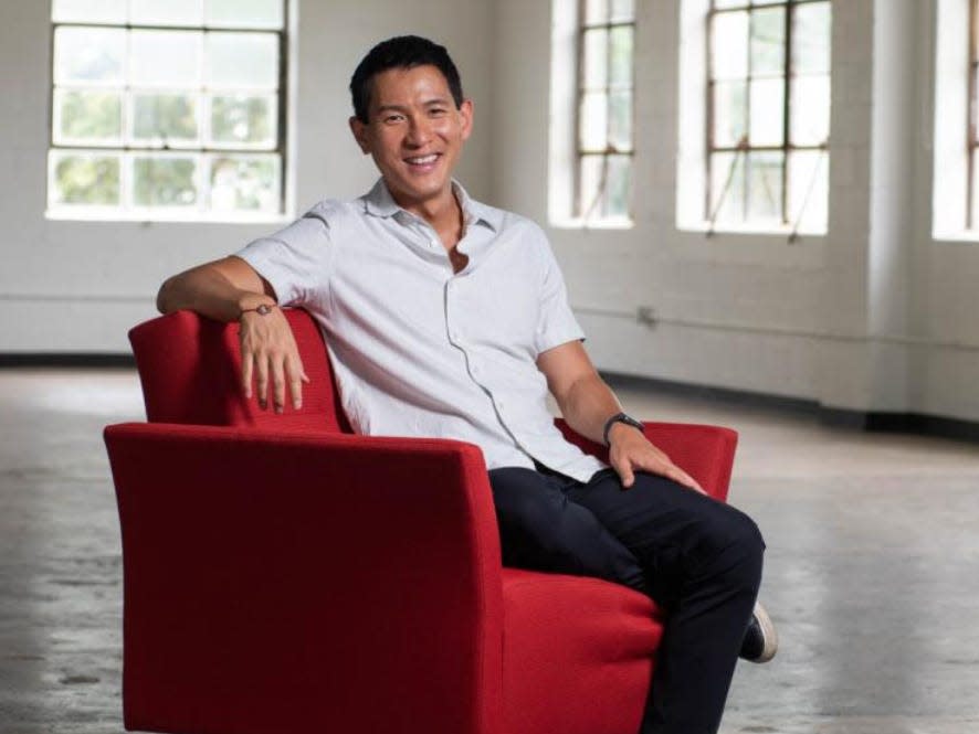 Chris Pan wearing a white collar shirt and black pants and sitting in a red chair in a warehouse space.