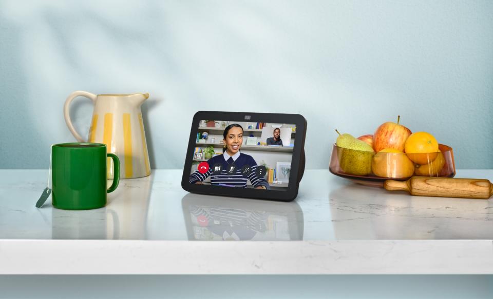 Amazon product photo of an updated Echo Show 8 smart speaker / display. It sits on a luxurious kitchen counter next to a mug, tea pot, and fruit basket. On its screen is a person smiling in a video chat.