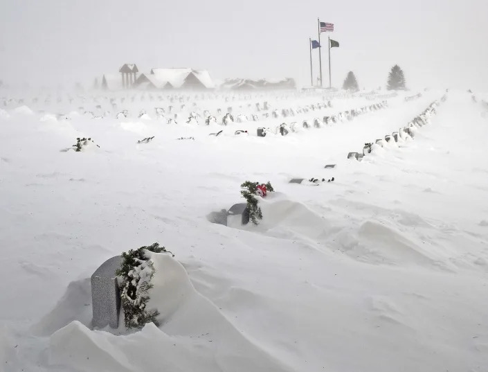 Rows of headstones, with Christmas wreaths, barely poke out of shrouds of snow, with a U.S. flag and two others flying in the far distance.