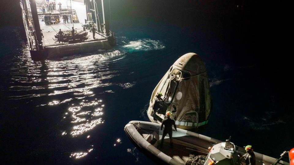 space capsule floats in a dark ocean with recovery crews in a small boat nearby.