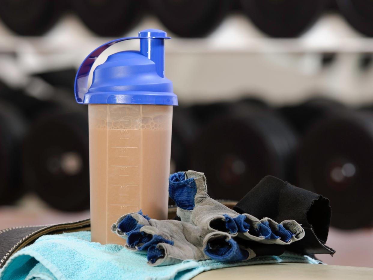 fitness bottle with protein shake sitting on a towel, lifting gloves near dumbbells