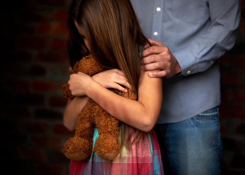 A little girl hugging her teddy bear while a man holds her shoulders. 