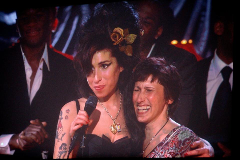 amy winehouse hugs her mother with one arm and holds a microphone on a stand in the other, both women smile as they stand on stage with a few men in suits in the background