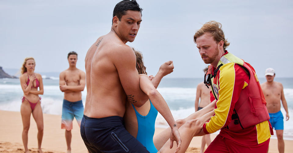 Home and Away extras stand on Palm Beach in swimwear, while the main actors carry out a surf rescue. 