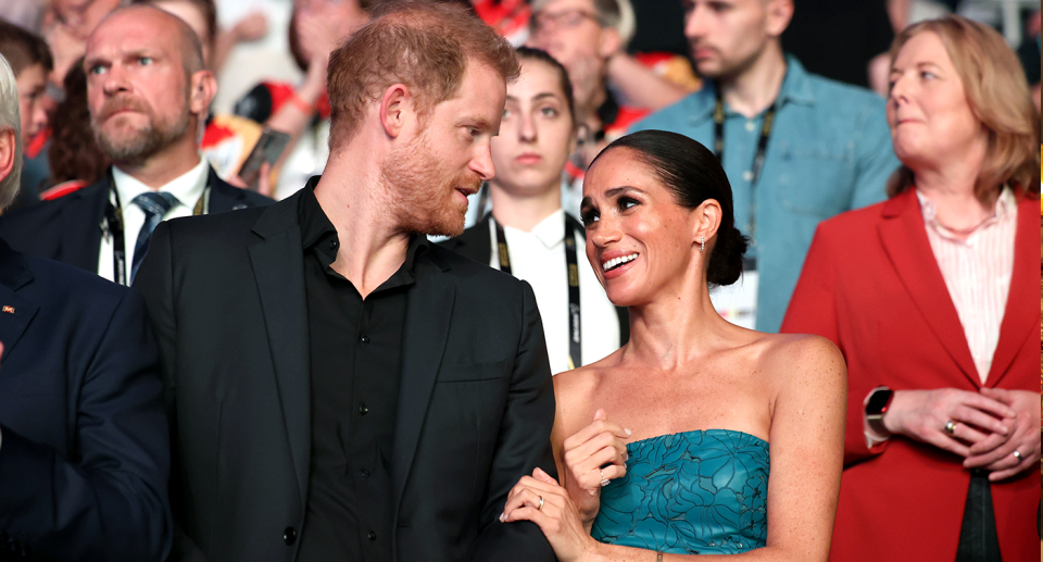 Prince Harry and Meghan Markle stare at one another in front of a crowd and Meghan places her hand on Harry's forearm. 