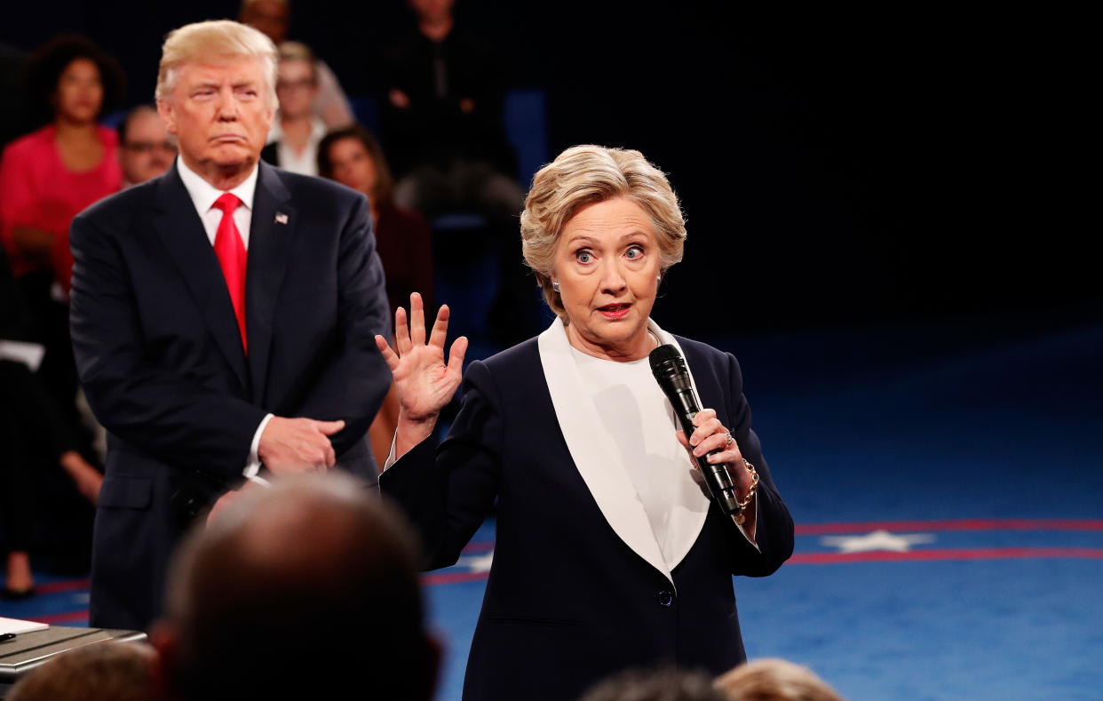 Hillary Clinton stands on a blue carpet, holding up her right hand with fingers extended while holding a microphone with her left, while Donald Trump glowers a few paces behind her.
