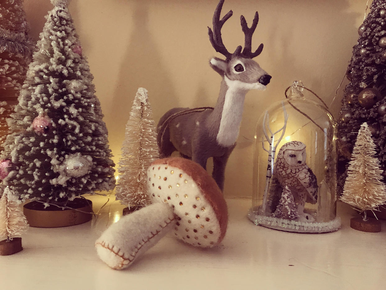 Close up of tiny, sparkling Christmas trees decorated with shiny silver and pink ornaments and glitter arranged neatly on a fireplace mantel. Tiny snow globe ornament with woodland owl figurine inside. Felt toadstool with glittering jewels. Reindeer. Festive holiday and celebration.